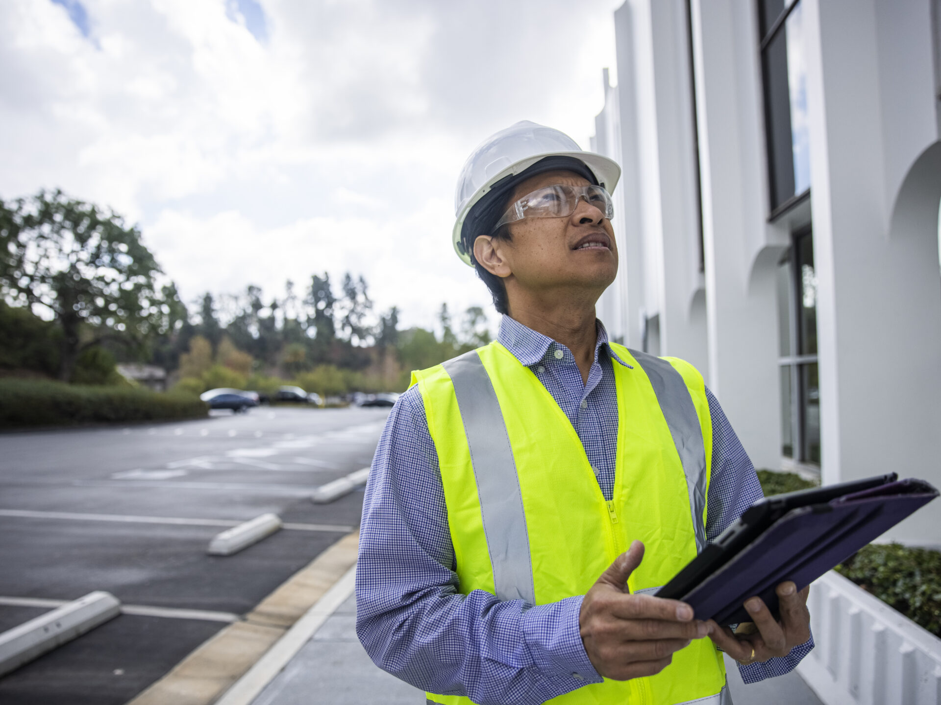 A mature Filipino man with a tablet and safety vest, helmet and glasses inspects a building and takes notes.