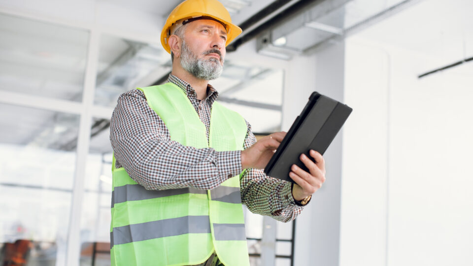 Mature bearded inspector in glasses and hardhat checking construction process with digital tablet. Male engineer using modern technology for work indoors.